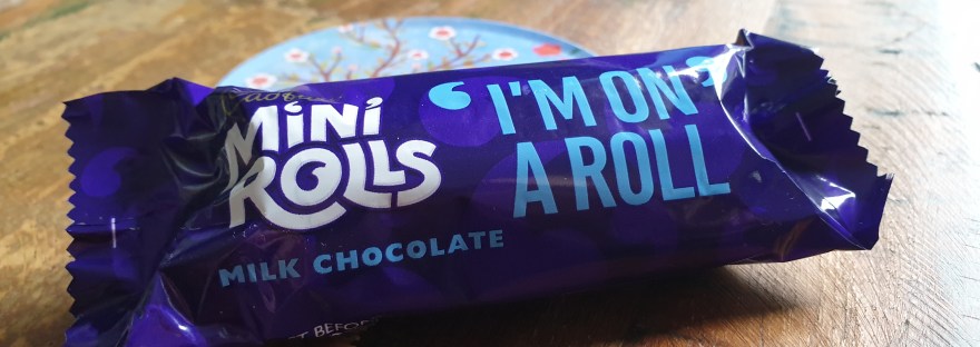Close up of a packaged Cadbury's Mini-Roll on a dark wooden table. Packaging features the phrase "I'm on a Roll"