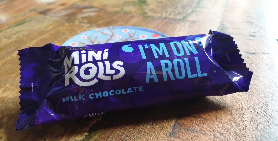 Close up of a packaged Cadbury's Mini-Roll on a dark wooden table. Packaging features the phrase "I'm on a Roll"