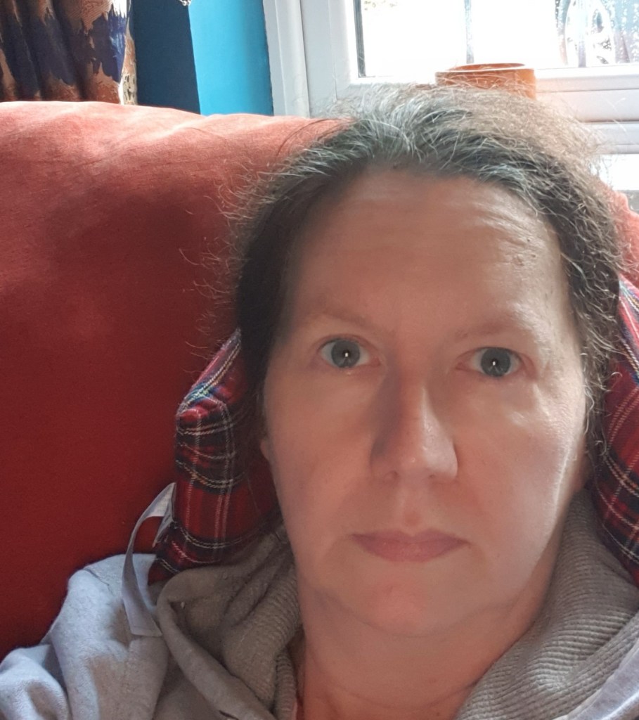 Me sitting upright on sofa looking at the camera with pained expression.  Wheat pack behind my head to provide relief